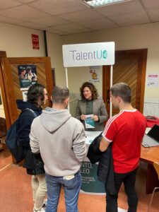 La ténica de Herramientas para el Empleo TalentUO ubidad en el stand informativo del hall mostrando un folleto del programa a tres alumnos de la Escuela 